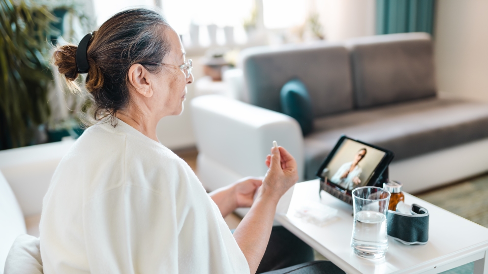 Woman in white robe sits on couch, video call on tablet, glass of water beside her.