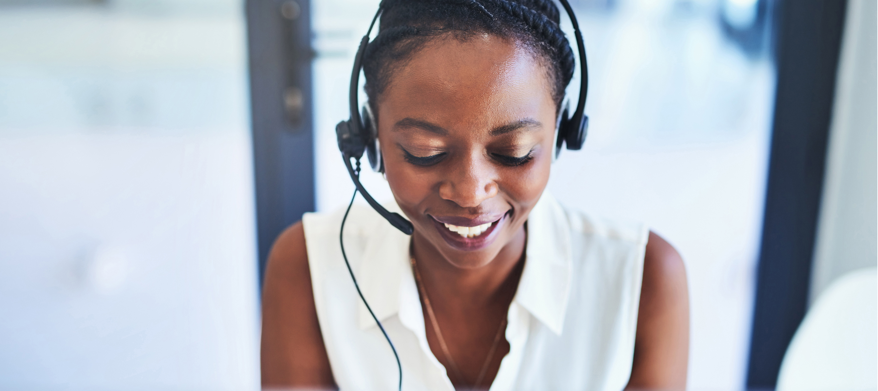 Smiling customer service agent wearing headset, focused on laptop screen in bright office setting.