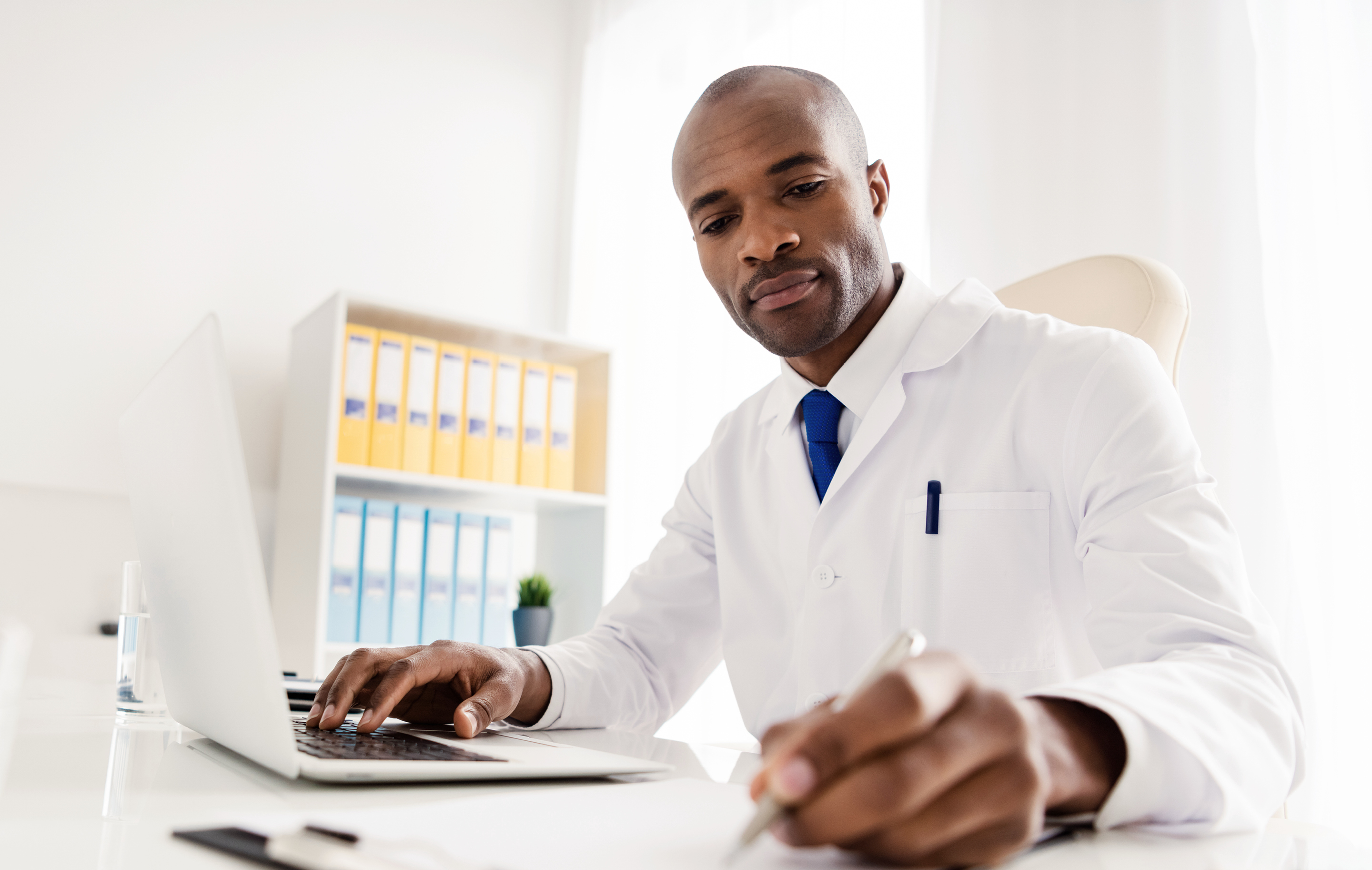 Healthcare professional in white coat typing on laptop at desk with office supplies nearby.