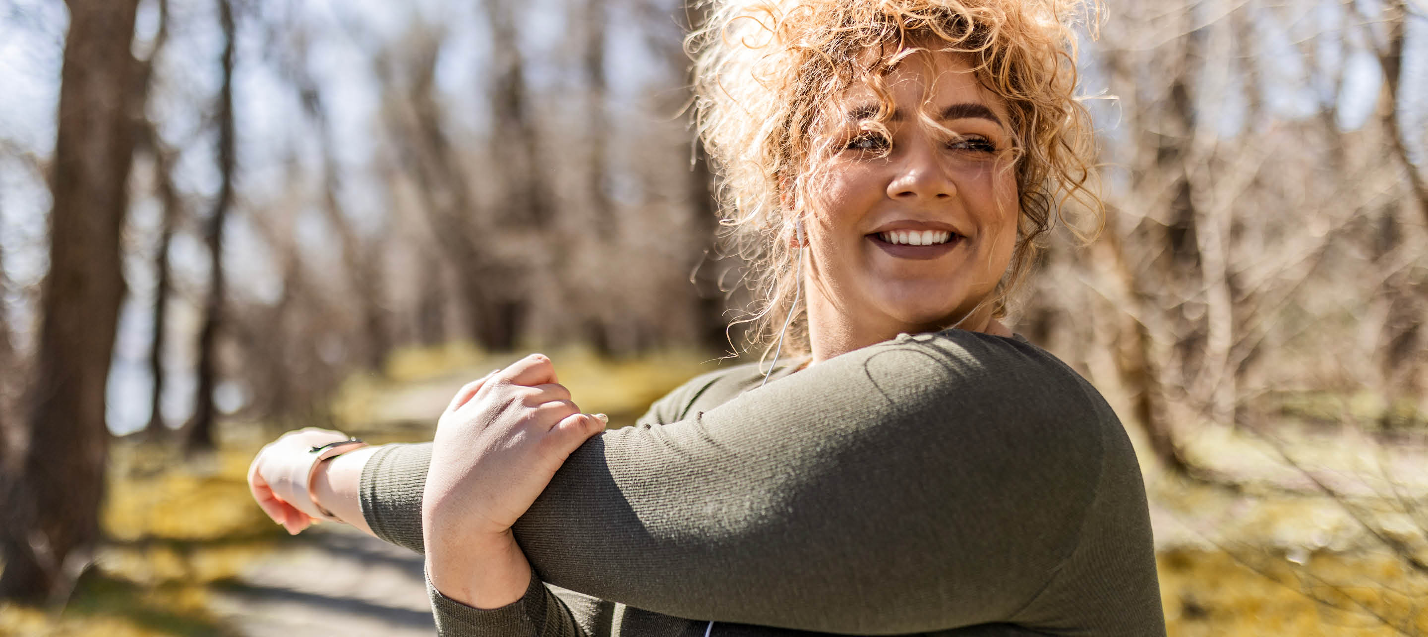 Woman smiles while stretching outdoors in a sunlit, wooded area wearing a dark green top.