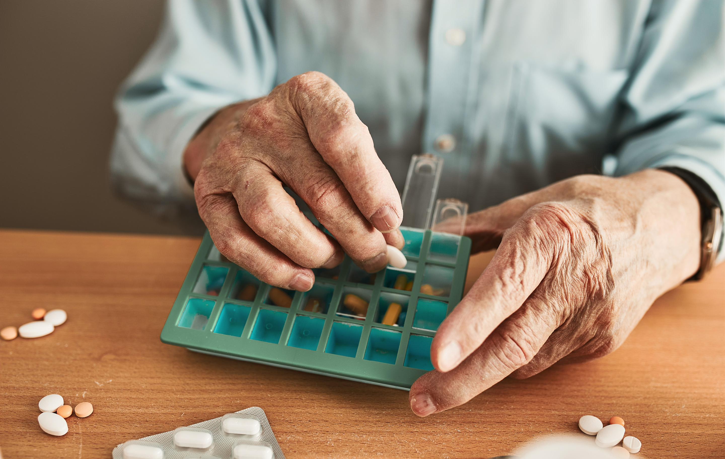 An elderly person carefully places pills into a blue weekly pill organizer on a wooden table.