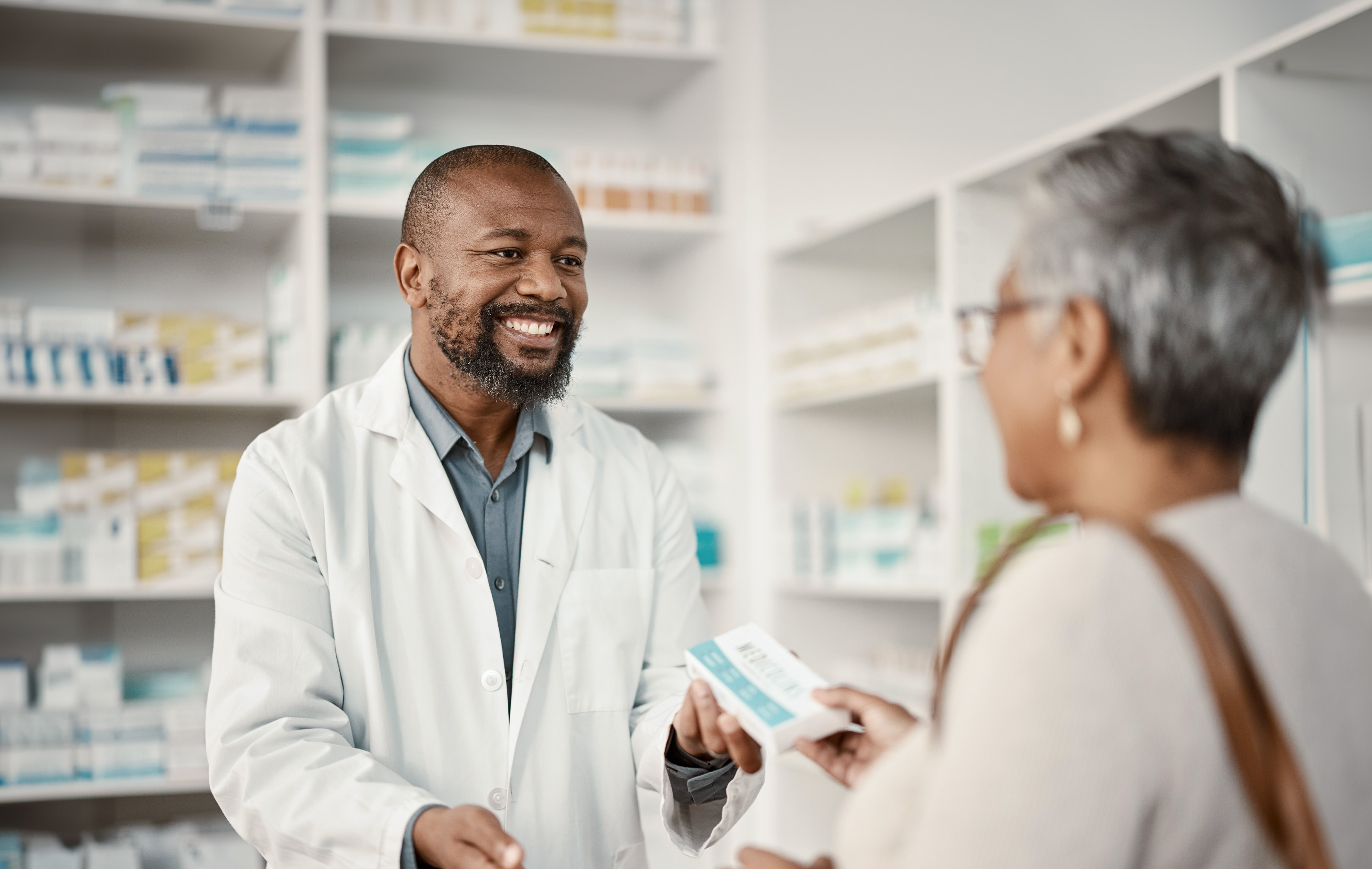 Pharmacist smiles while handing medication to a customer in a well-lit pharmacy.