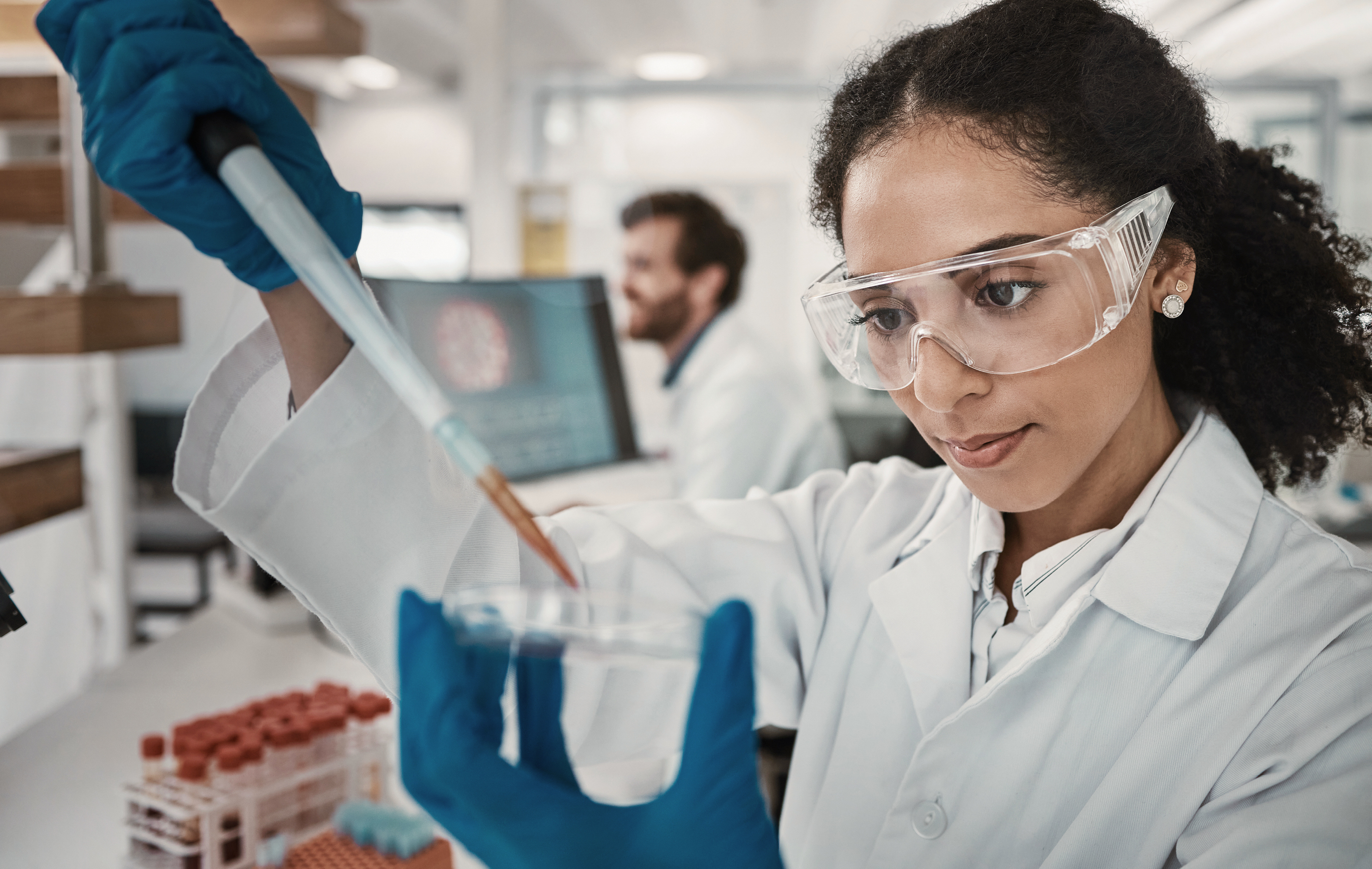 Scientist in lab coat and goggles carefully transfers liquid with pipette into test tube.