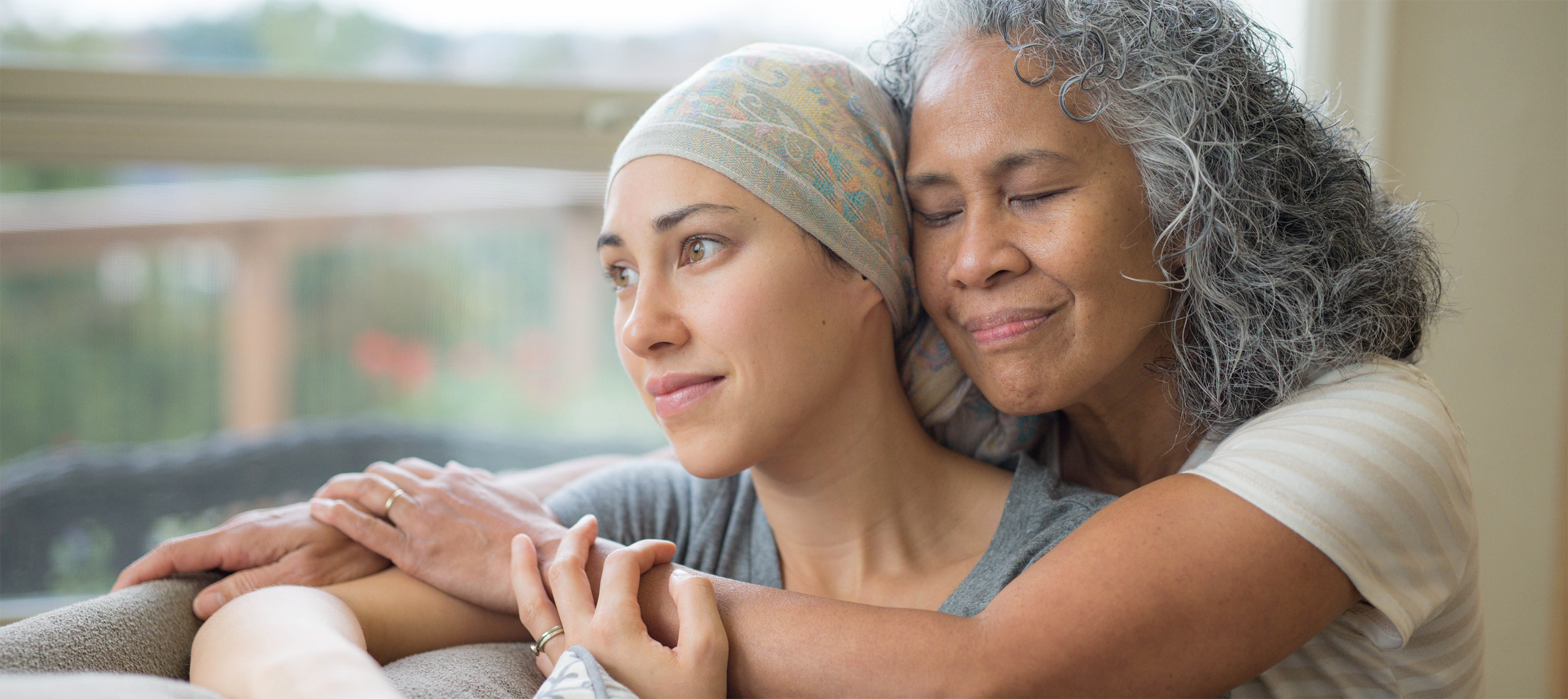 Two women embrace warmly by a window one with a headscarf the other with long gray hair