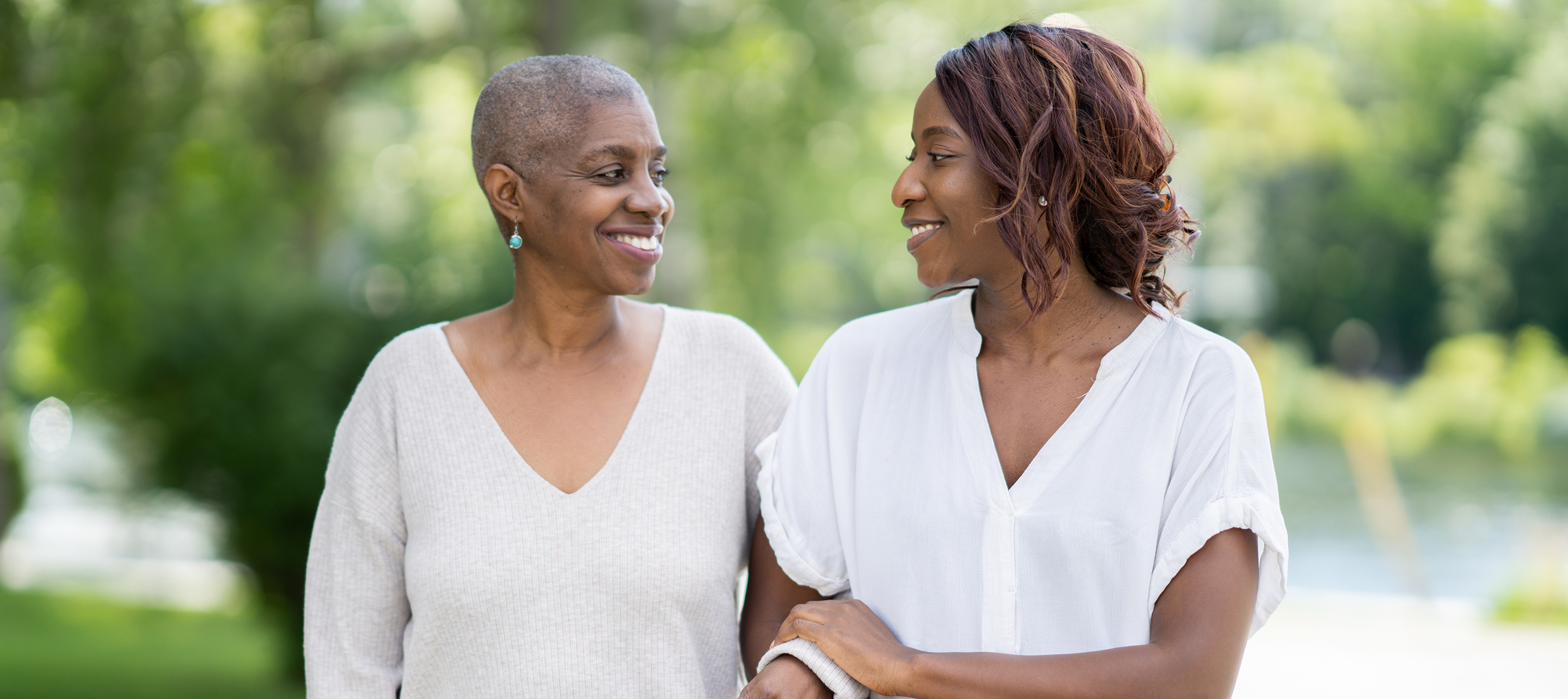 Two women in white shirts smiling and walking together in a sunlit park.
