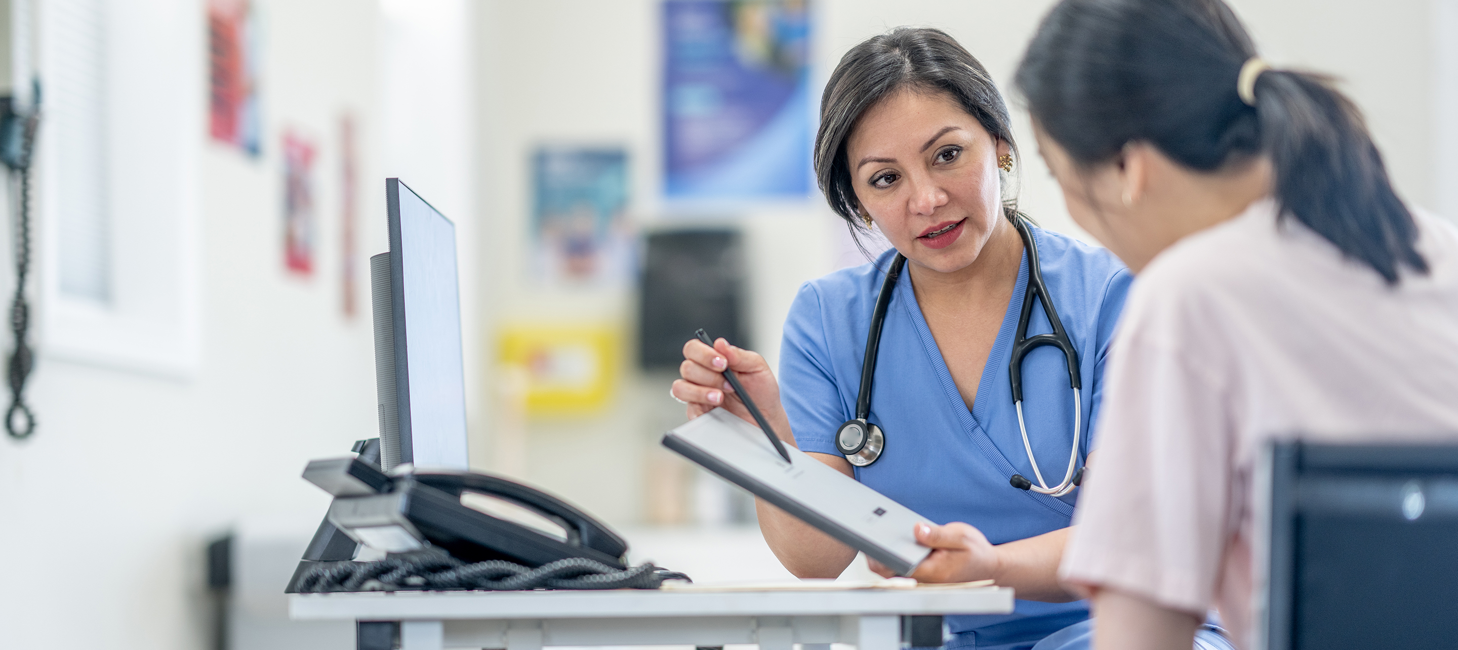 Medical professional in blue scrubs explains something to a patient using a tablet at a clinic desk.