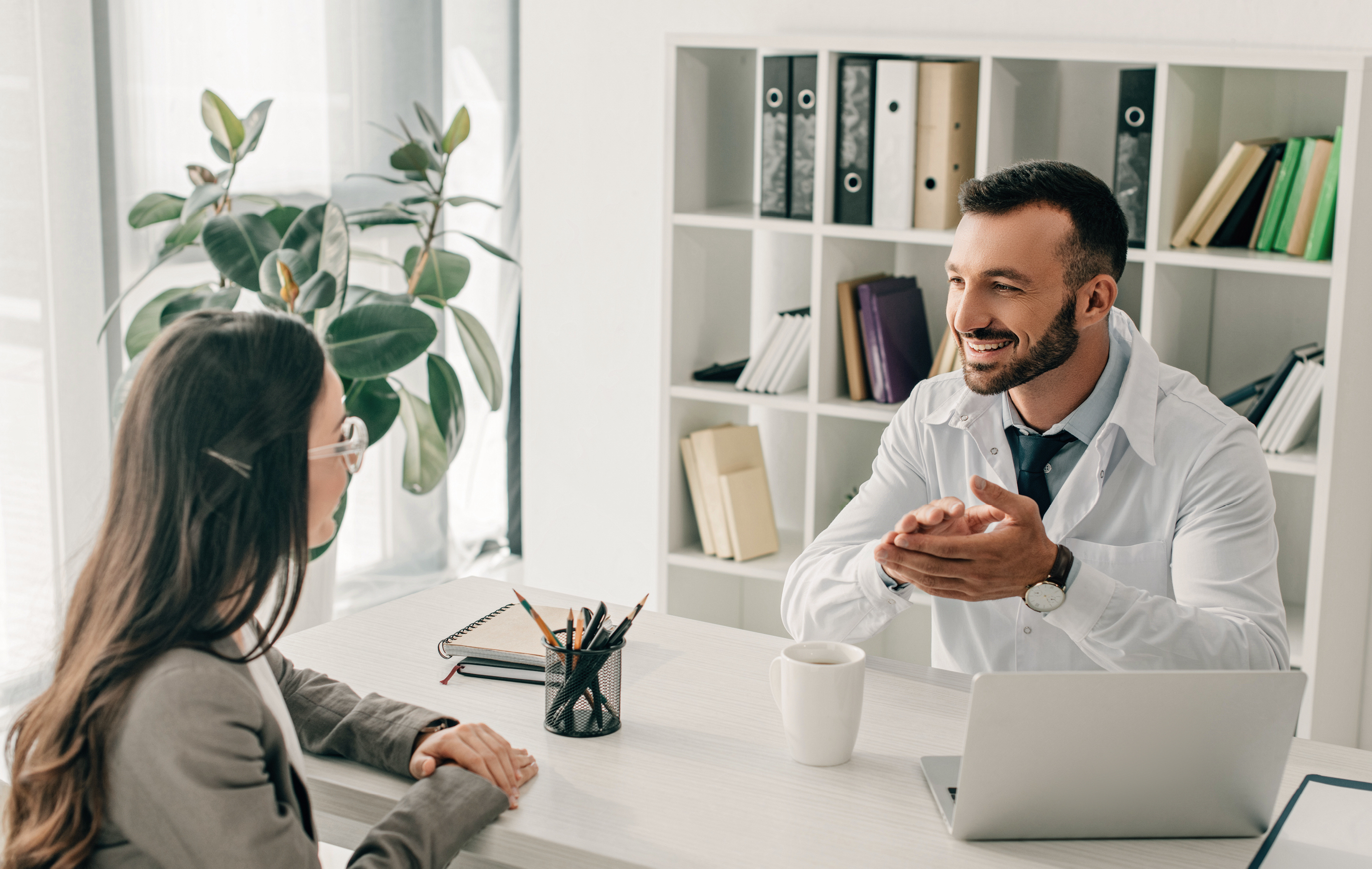 A smiling male doctor in a white coat talks to a female patient in a bright, organized office.