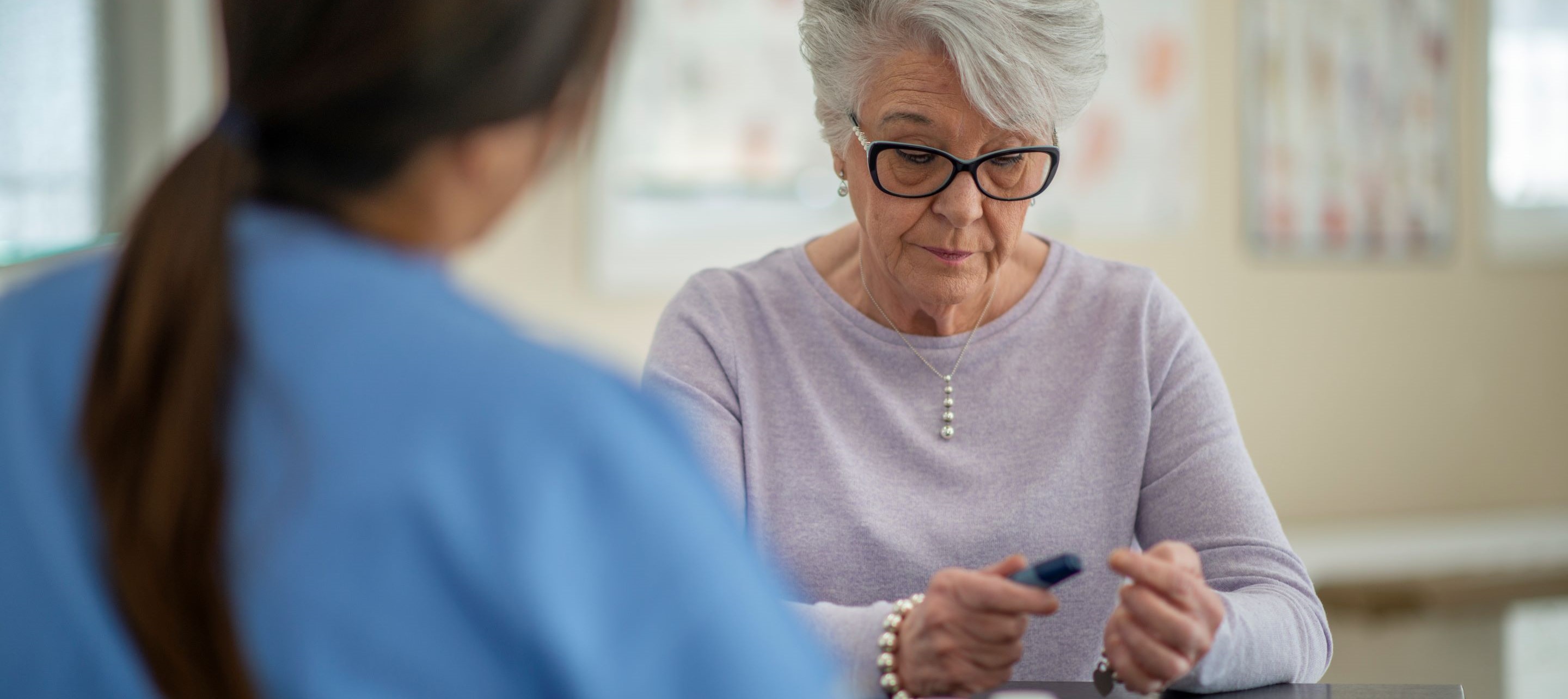 Senior woman with glasses checks blood sugar level with healthcare provider in clinical setting.