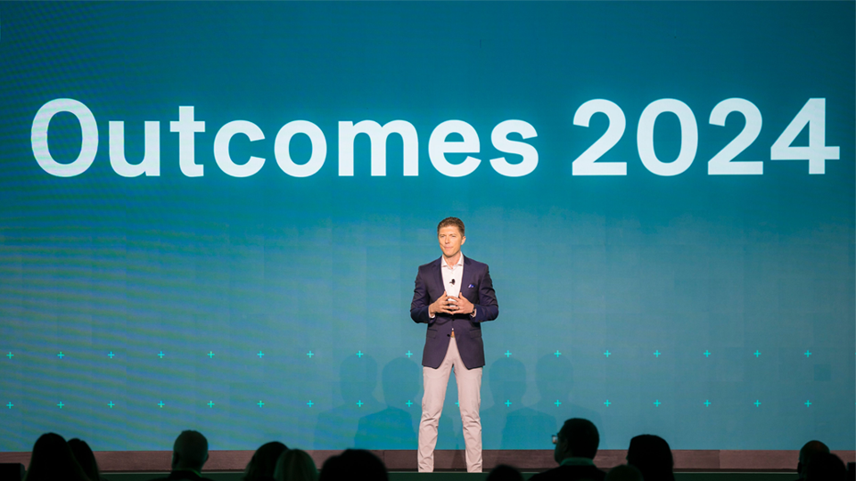 Adam Kautzner on stage before audience with Outcomes 2024 projected behind them on blue backdrop