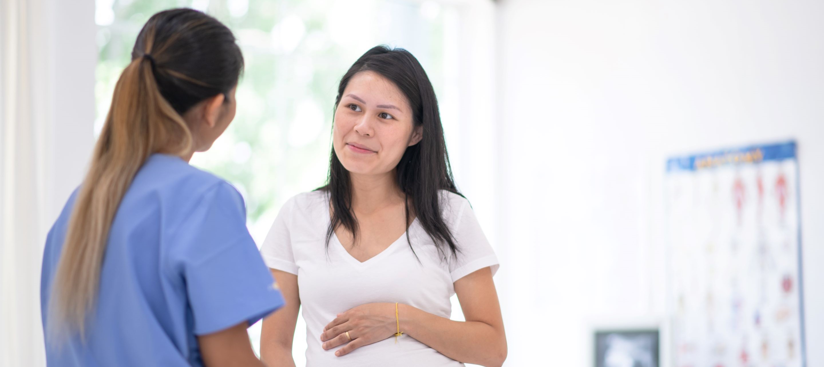 A healthcare provider in blue scrubs speaks with a pregnant patient in a white shirt
