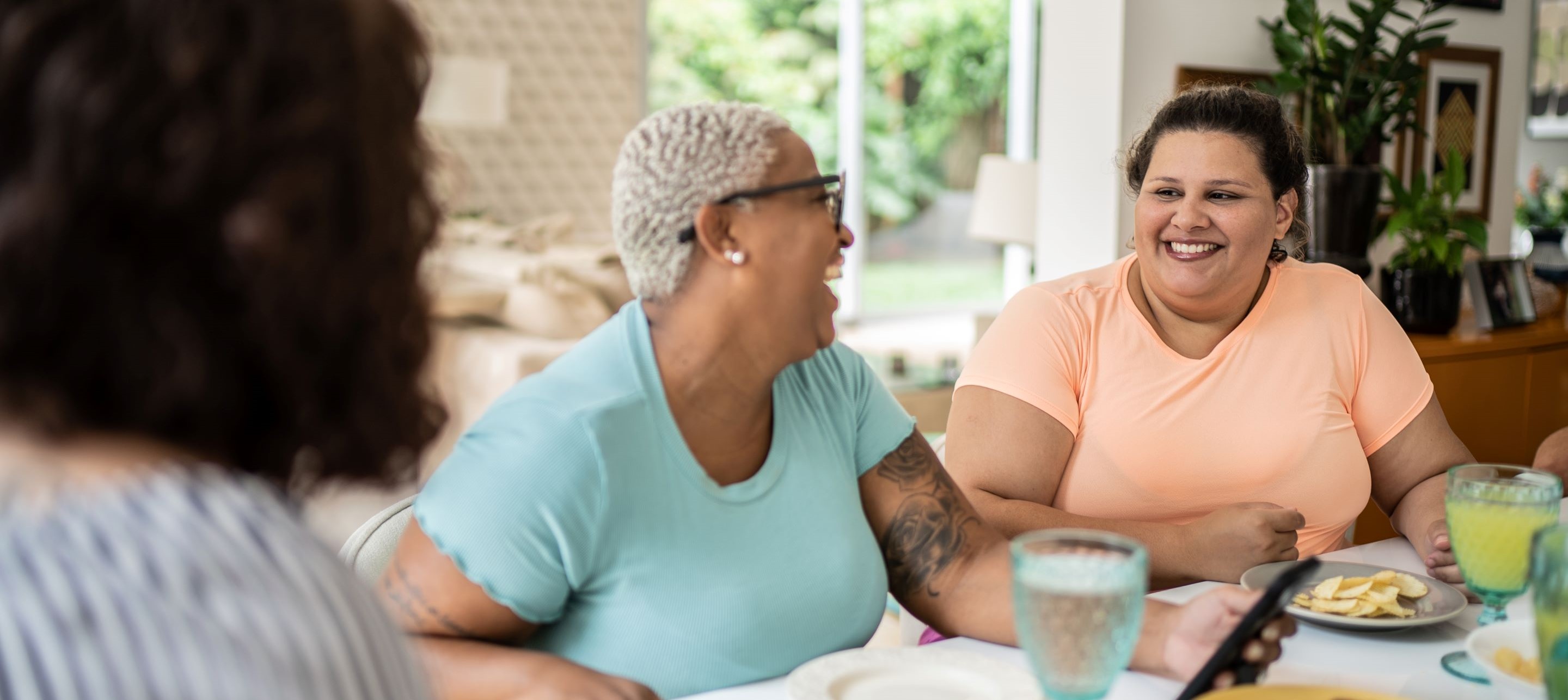 Three women share a warm laugh over a meal in a bright, cozy living room.