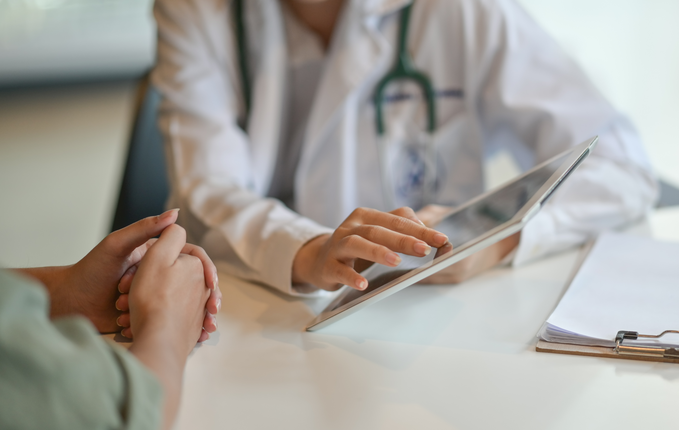 Doctor in white coat with stethoscope uses tablet with patient seated at desk during consultation.