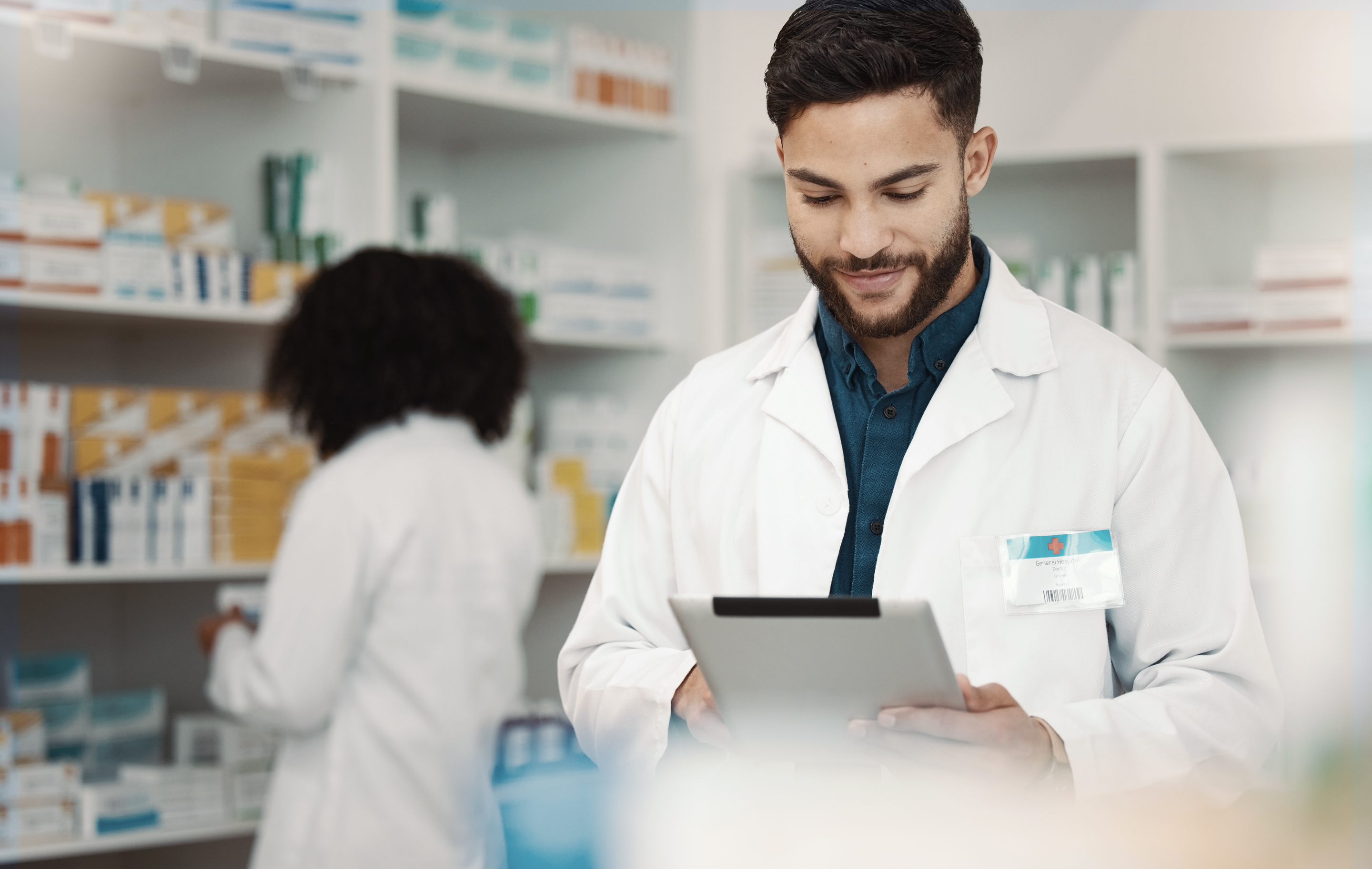 Pharmacist smiling while using tablet in well-lit pharmacy with shelves and another staff member in background.