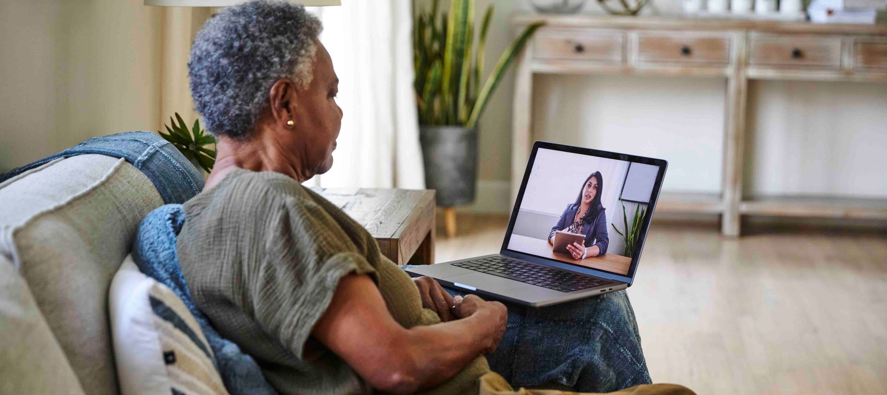Senior woman sits on couch using laptop for video call in bright, cozy home setting