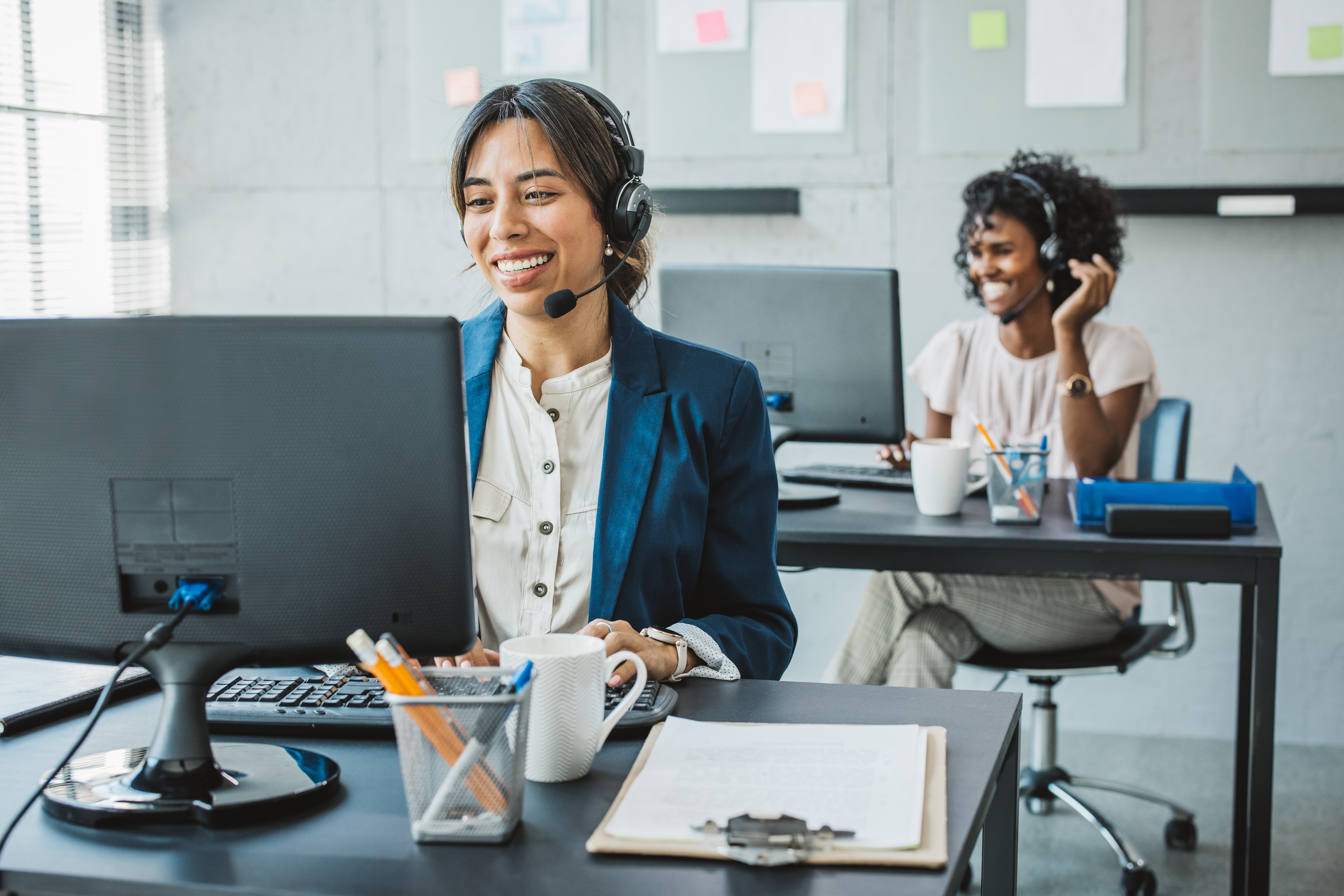 Two smiling agents in a modern office, wearing headsets, working at desks with computers.