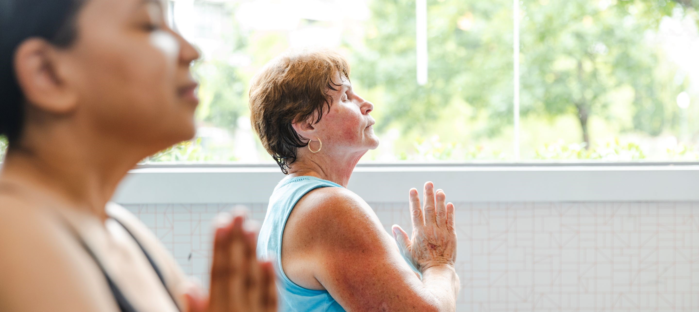 Two women practice mindfulness in a sunlit room with large windows.