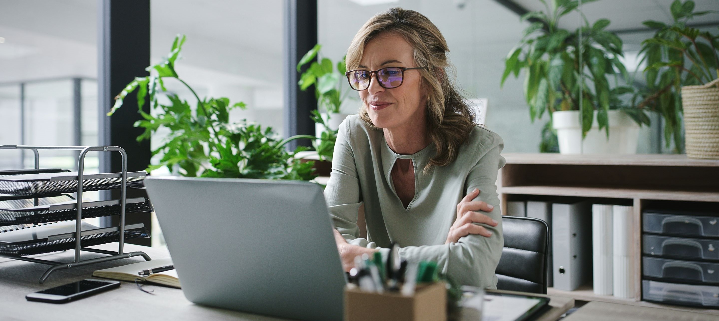 Woman with glasses smiles while working on laptop in bright, plant-filled office space.