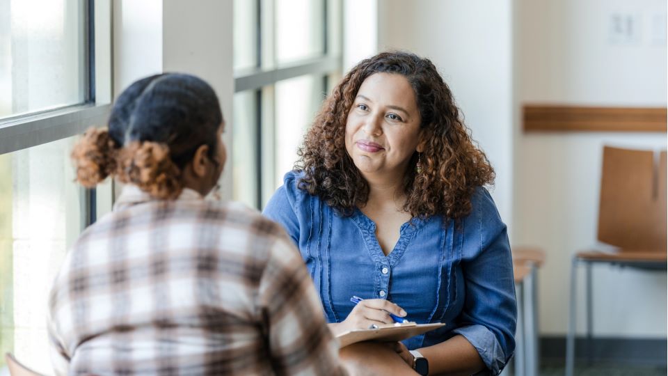 Woman with curly hair and blue shirt observing another woman holding a clipboard.