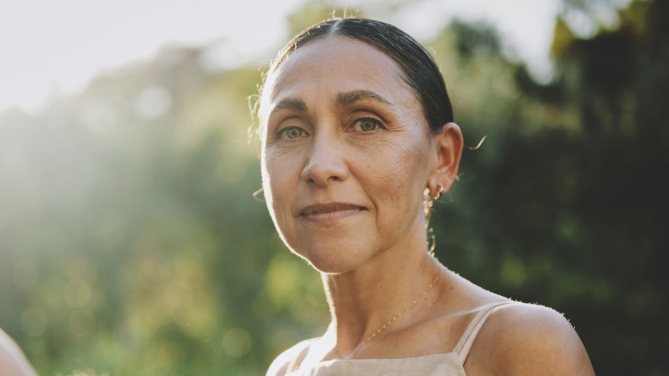 Woman with dark hair, smiling slightly, wearing earrings and a cream-colored sleeveless top.