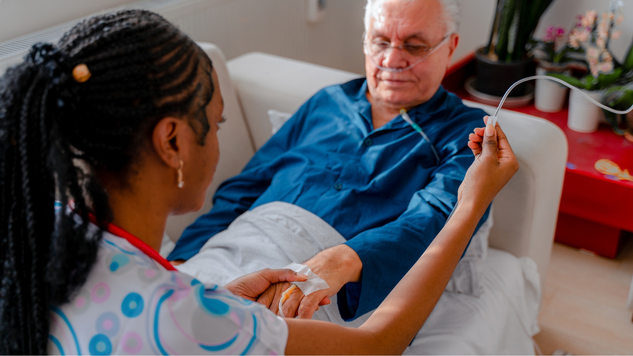 Older man on bed in blue shirt, woman with braided hair, holding his hand.