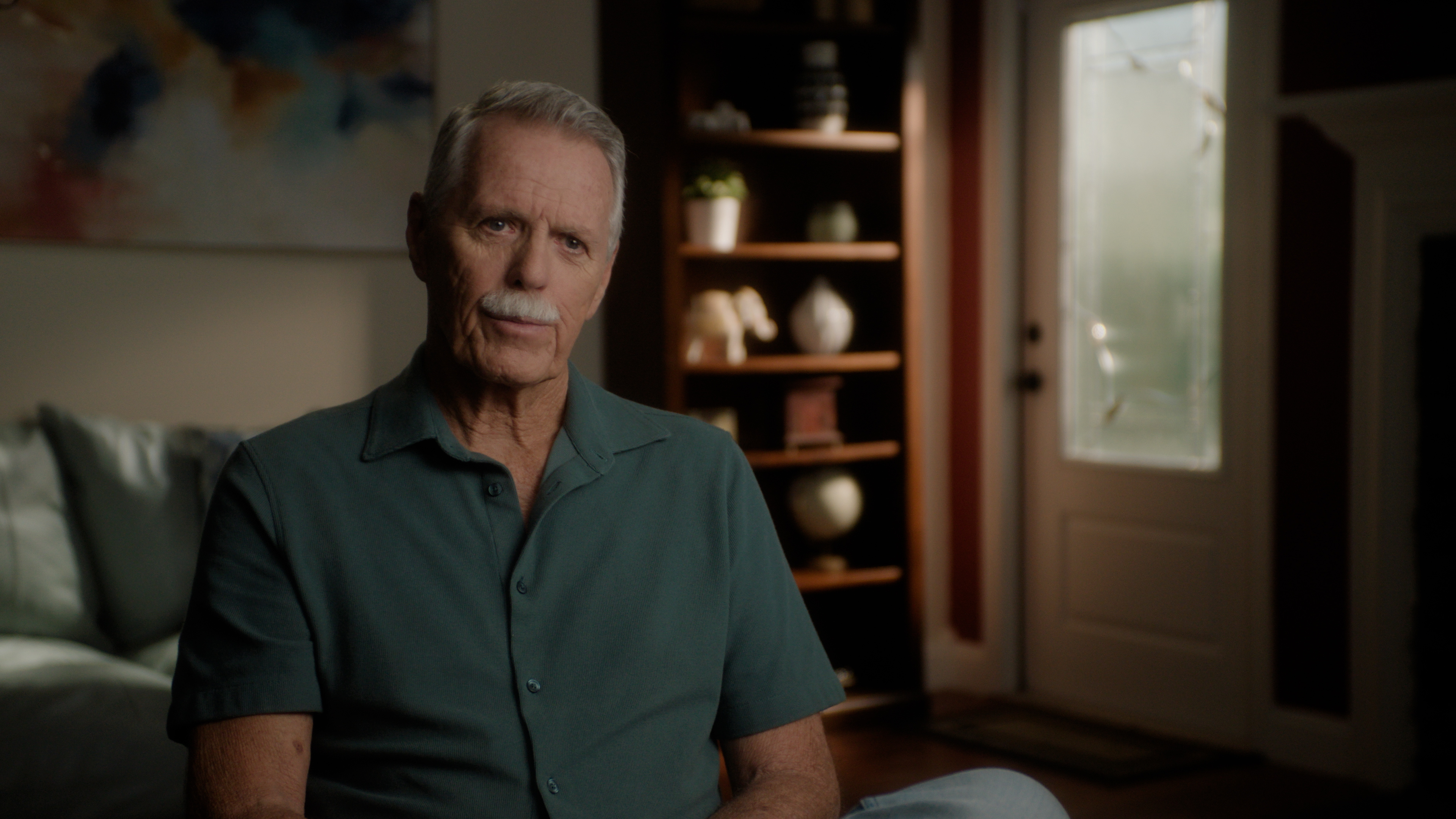 An elderly man with gray hair and a mustache sitting on a couch in a room with a bookcase.