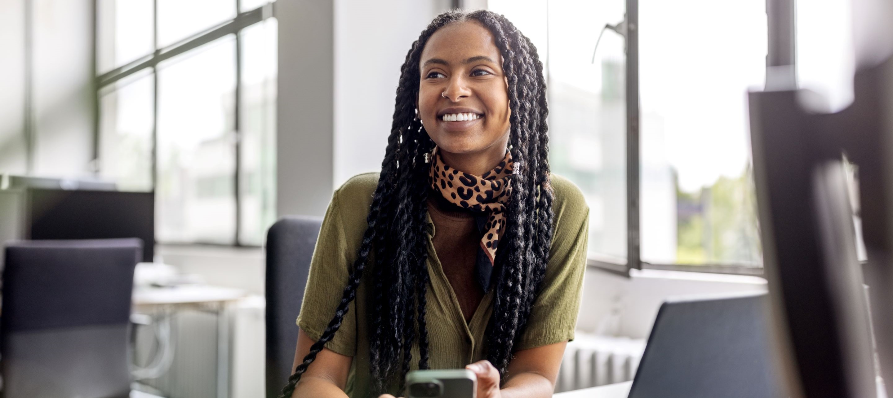 A woman in a green shirt with long braids, wearing a leopard print scarf and seated at a desk next to a laptop