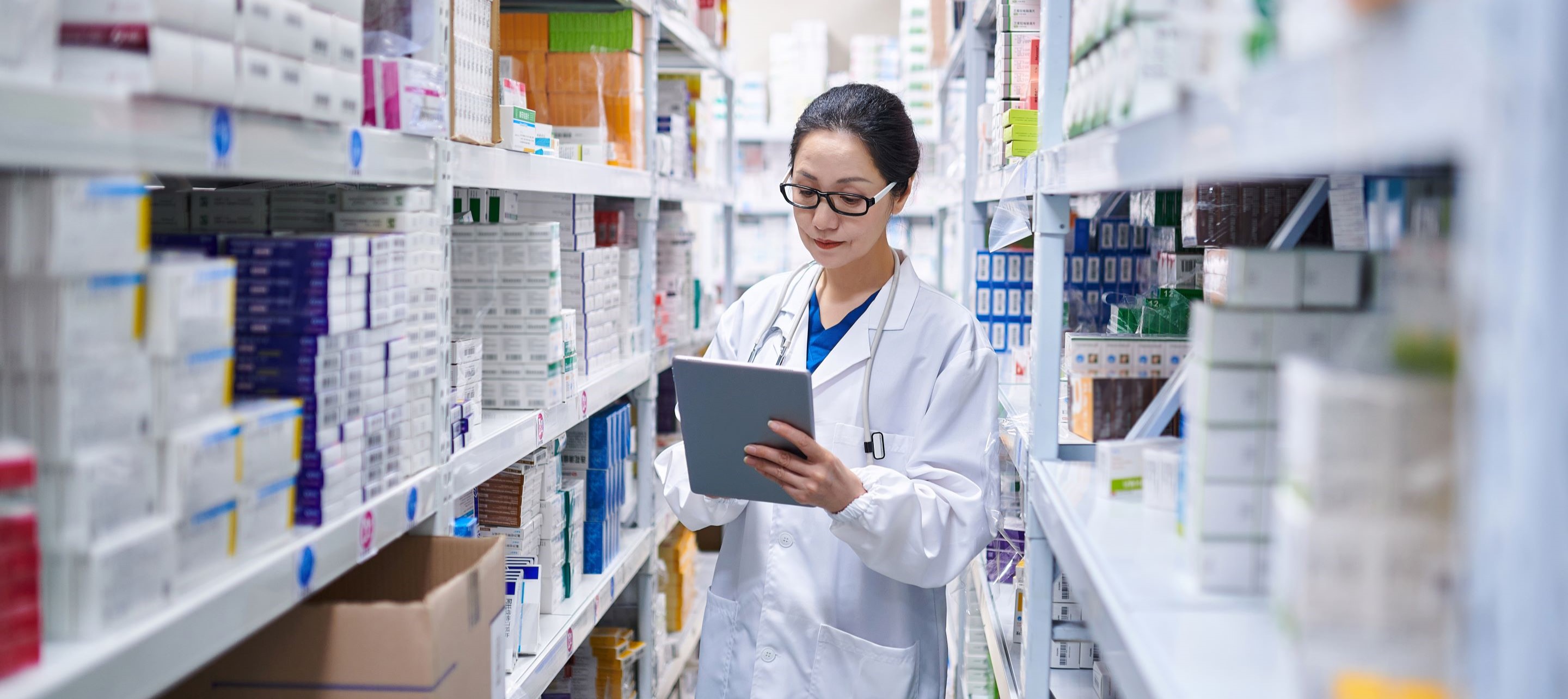 Pharmacist in white coat and glasses uses tablet in brightly lit pharmacy aisle.