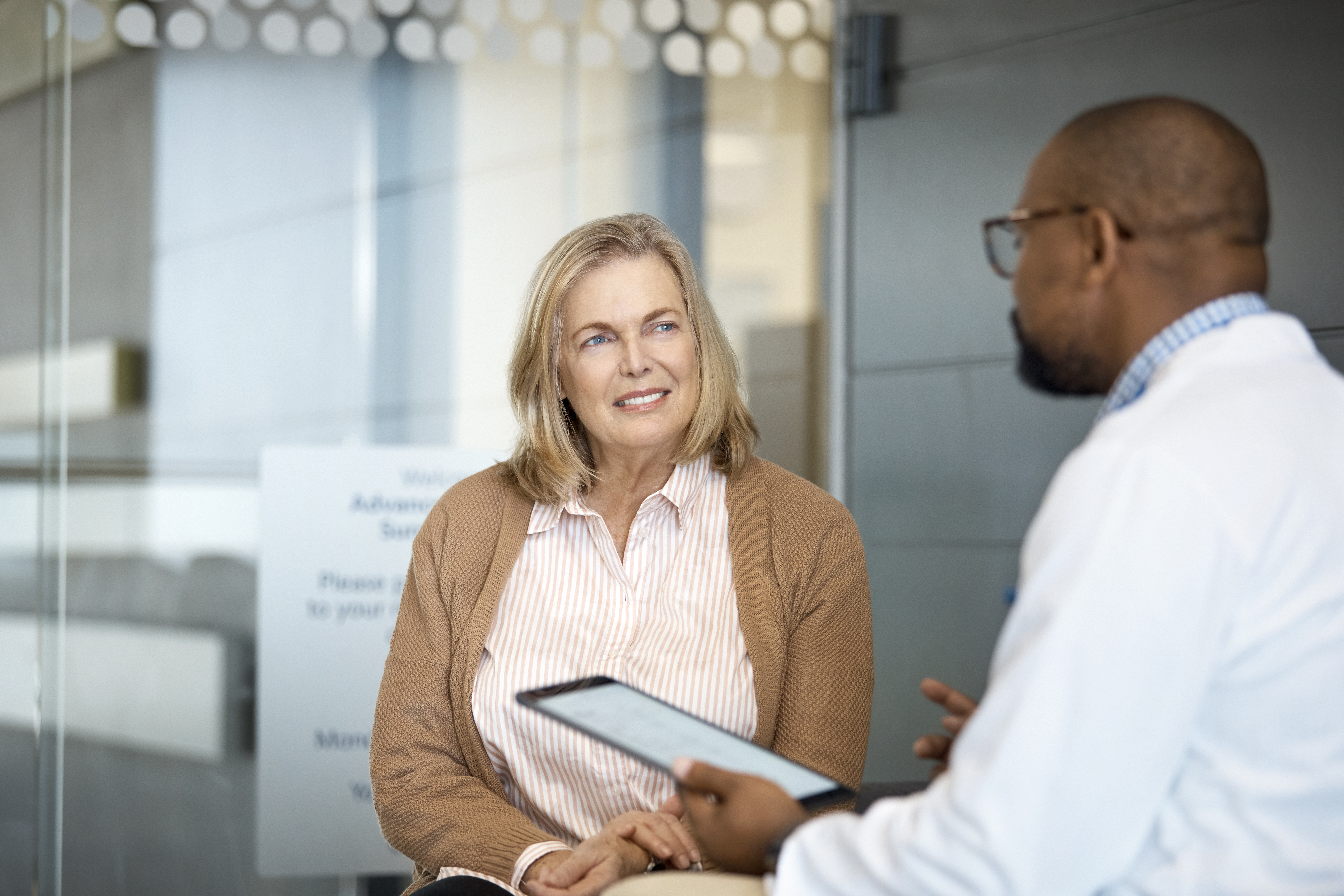 A smiling woman sits with a doctor in a clinic, who holds a tablet as they discuss health.