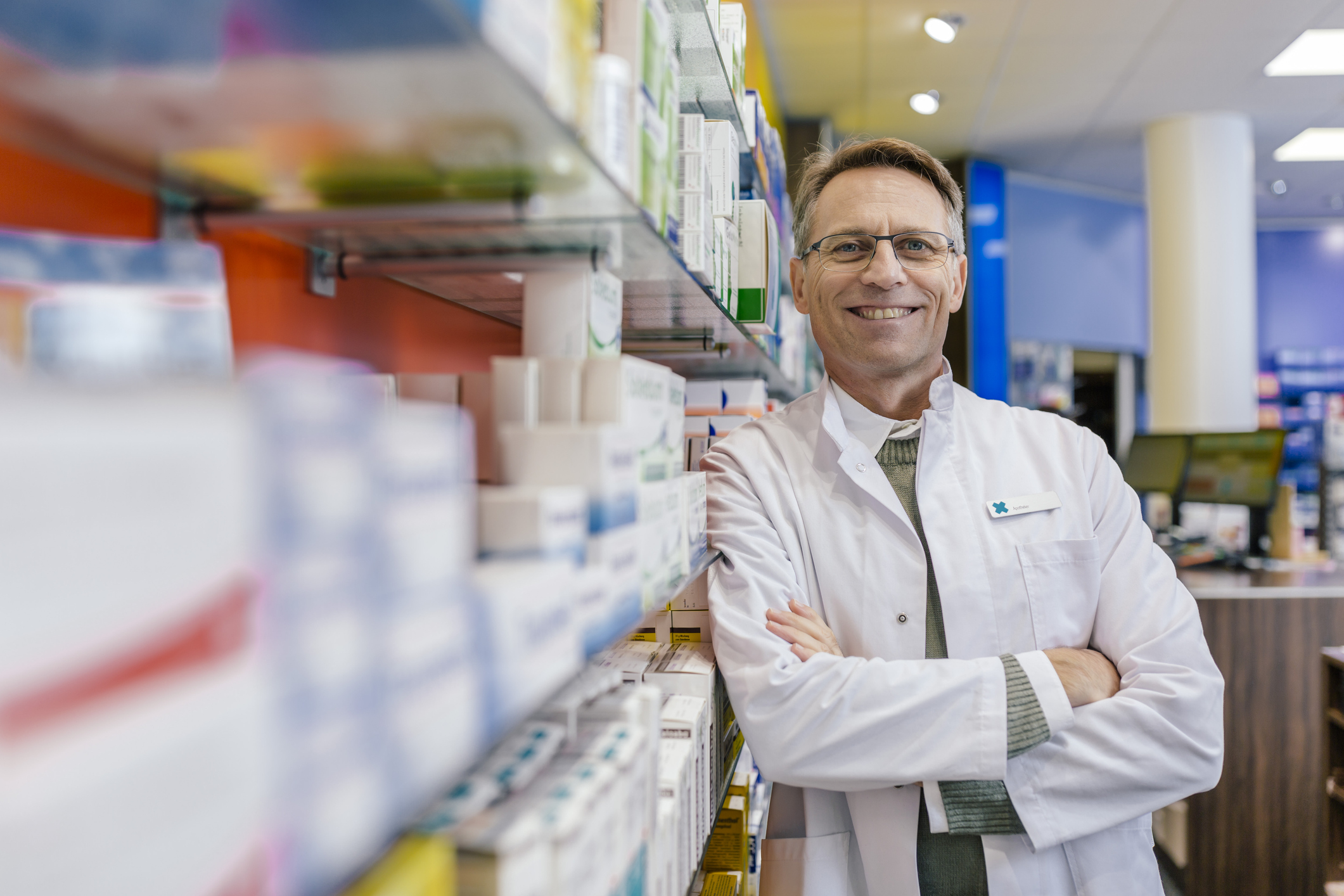 Pharmacist smiling, looking directly at camera and leaning on wall of prescriptions