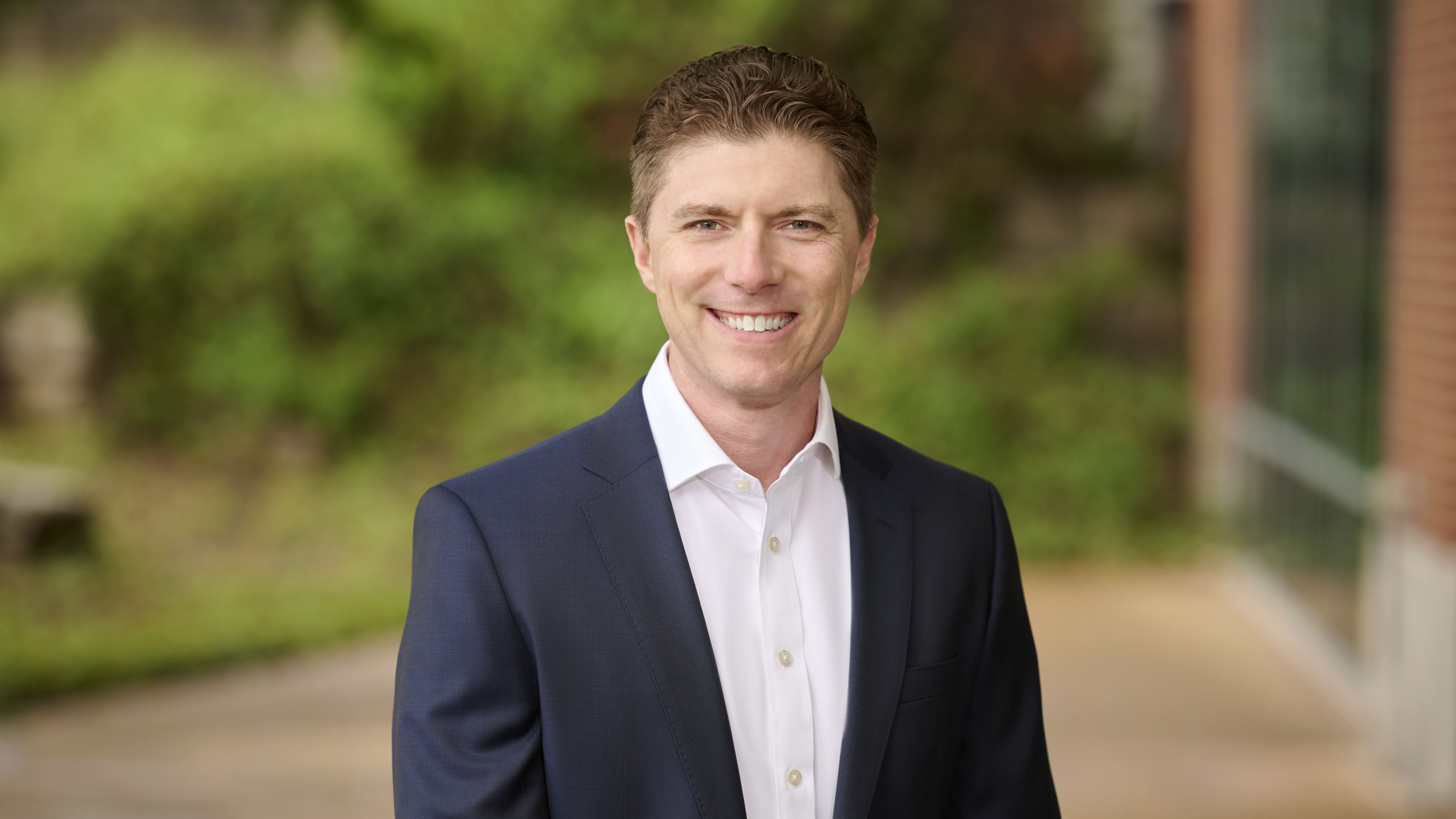 Adam Kautzner smiles at camera, wearing a suit with no tie