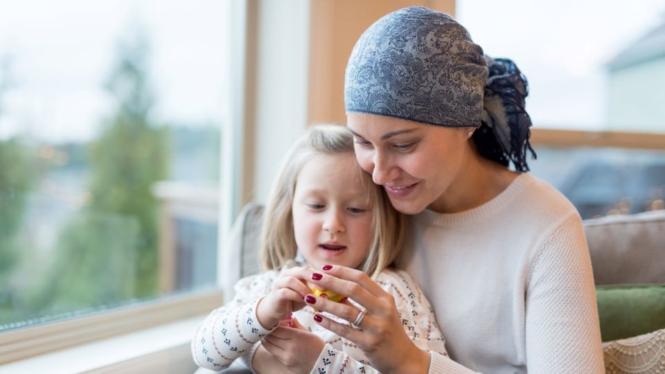 Adult wearing a patterned head scarf seated indoors near a window, holding and looking at a small object together with a young child. The adult and child are close together on a couch, with natural light coming through the window and a softly blurred outdoor background.