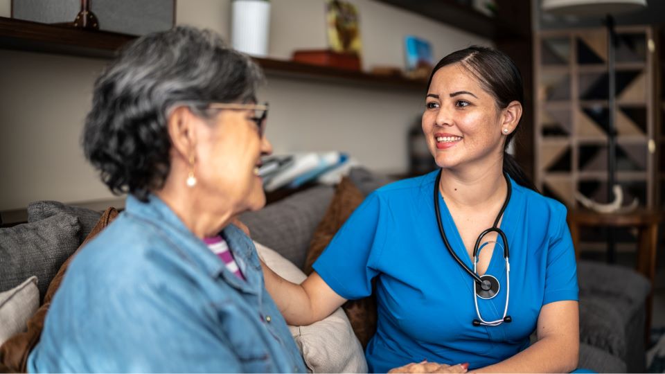 Nurse in blue scrubs with a stethoscope sits beside an older adult on a couch, offering in‑home care.