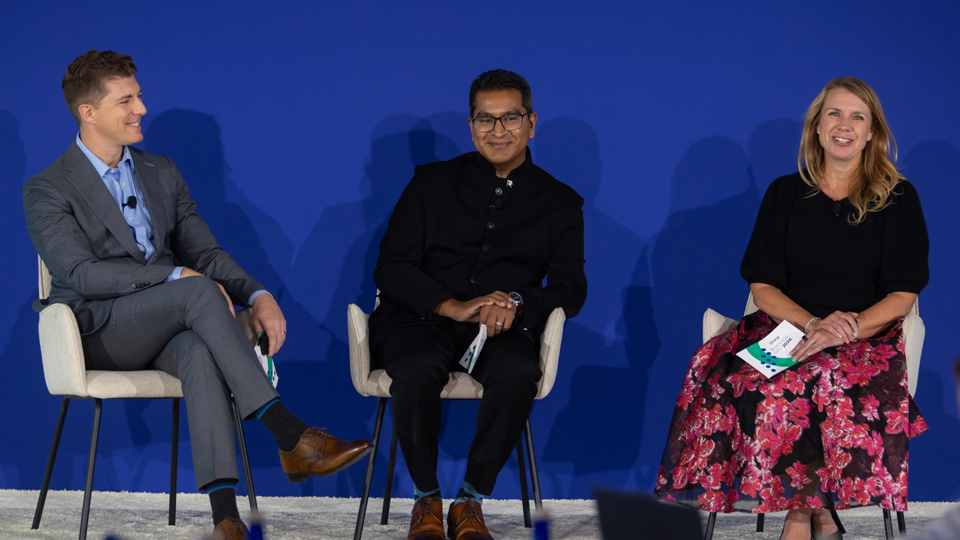 Three panelists sit on stage in front of a blue backdrop during a discussion. From left to right, Adam Kauzner, AJ Ally, and Alysha Fluno are seated in white chairs holding event materials while participating in the conversation.