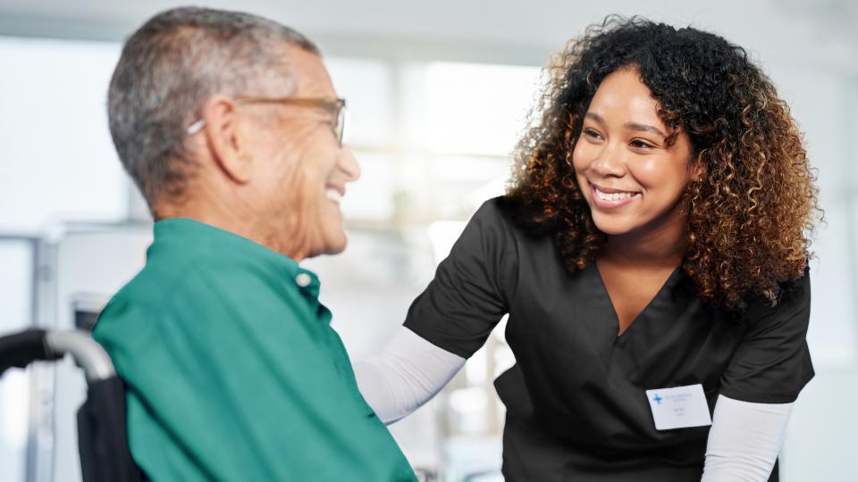 A nurse is smiling at an elder man sitting in a wheelchair.