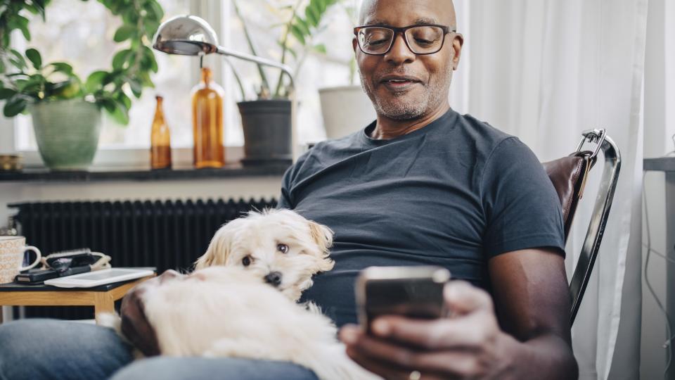 A man sits at home with a small dog and uses a smartphone