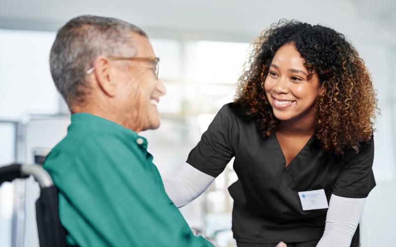 A nurse is smiling at an elder man sitting in a wheelchair.