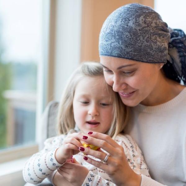 Adult wearing a patterned head scarf seated indoors near a window, holding and looking at a small object together with a young child. The adult and child are close together on a couch, with natural light coming through the window and a softly blurred outdoor background.