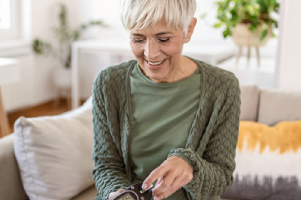 Smiling woman with short white hair checks blood sugar meter while seated on couch in bright living room.